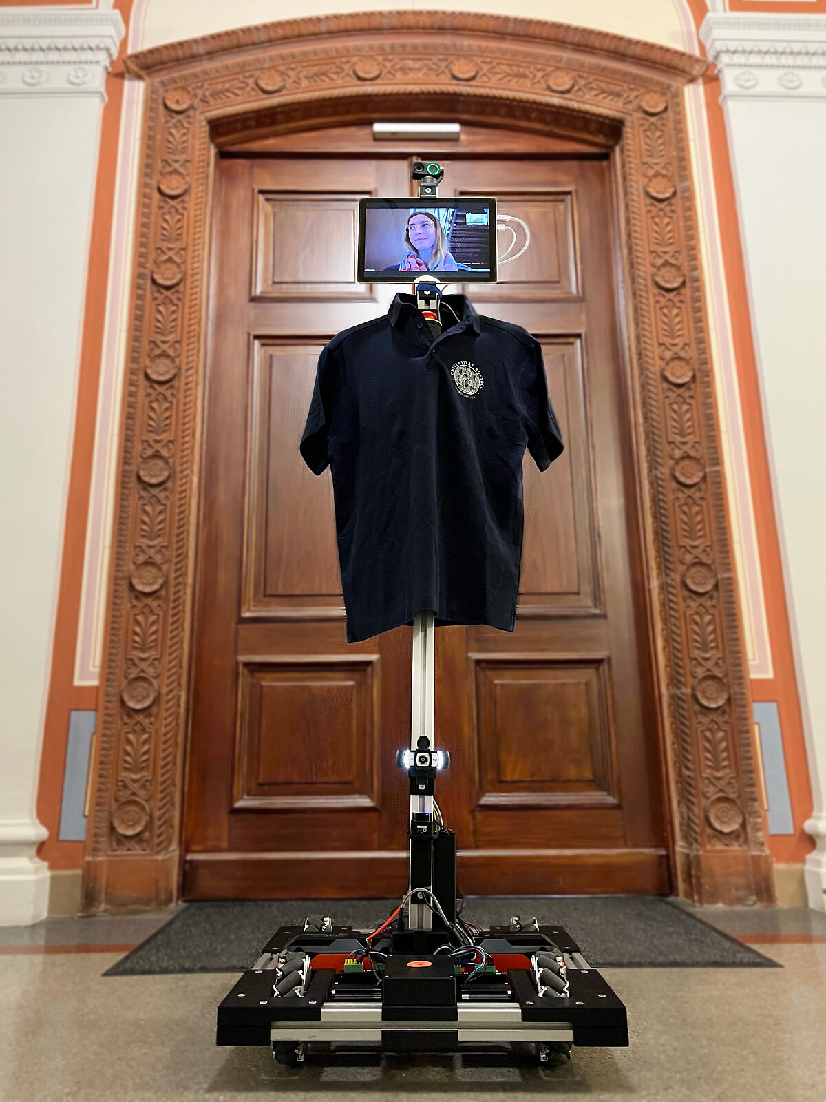 Das rechteckige Bild zeigt ein Foto des Telepräsenzroboters, das leicht aus der Froschperspektive aufgenommen wurde. Der Roboter befindet sich im Flur des Hauptgebäudes der Universität Rostock. Im Hintergrund ist eine große Tür zu sehen. Der Telepräsenzroboter ist mit einem blauen T-Shirt der Universität bekleidet. Auf seinem Bildschirm ist eine studentische Hilfskraft zu sehen, die leicht lächelt und ihren Blick nach links aus dem Bild richtet.